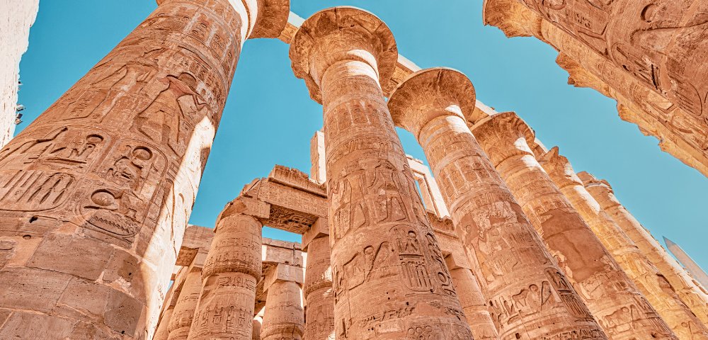 Ancient Egyptian stone columns with intricate hieroglyphics stand under a clear blue sky, conveying a sense of awe and historical grandeur.