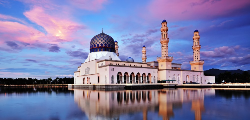 A grand mosque with a striking blue dome and four tall minarets is reflected in calm water at sunset, under a sky painted with pink and purple hues.