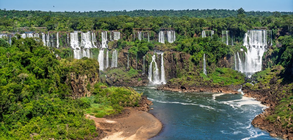 Lush green landscape with multiple waterfalls cascading into a river, surrounded by dense forest, evokes a serene and majestic natural scene.