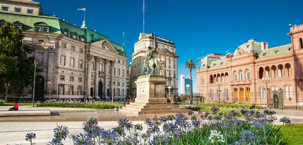 Historic square with grand neoclassical buildings and a central equestrian statue. Bright blue sky, vibrant flowers, and a few palm trees add charm.