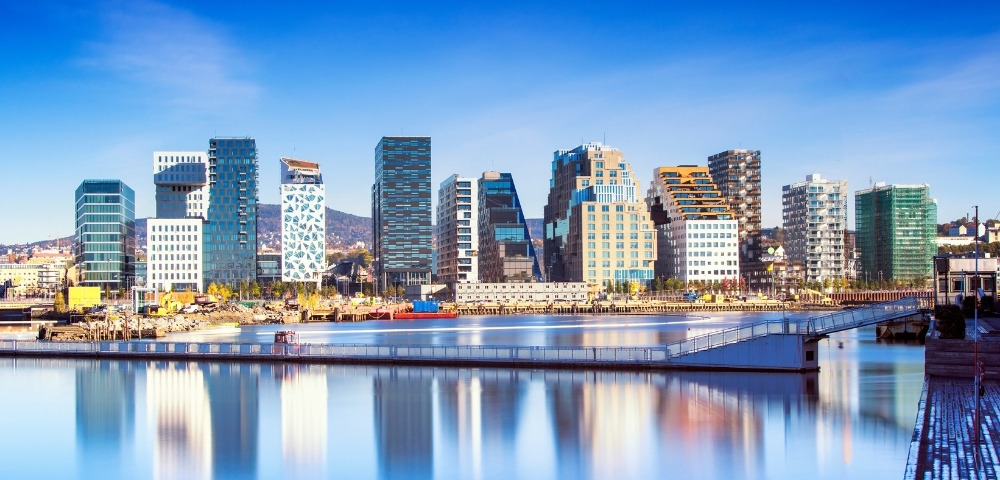 Panoramic view of Oslo's modern skyline with diverse, angular skyscrapers reflecting in a tranquil harbor under a clear blue sky, conveying urban serenity.