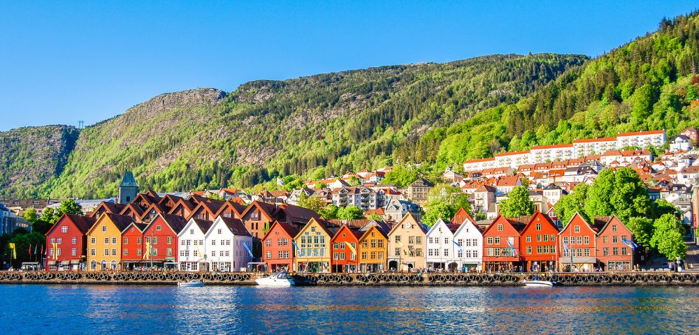 Colorful wooden houses line a waterfront in front of a lush green hillside under a clear blue sky, creating a vibrant and serene scene.