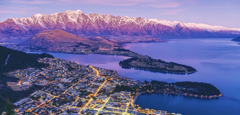 A vibrant cityscape at dusk, with sparkling lights, lies near a serene lake, framed by snow-capped mountains under a soft purple twilight sky.
