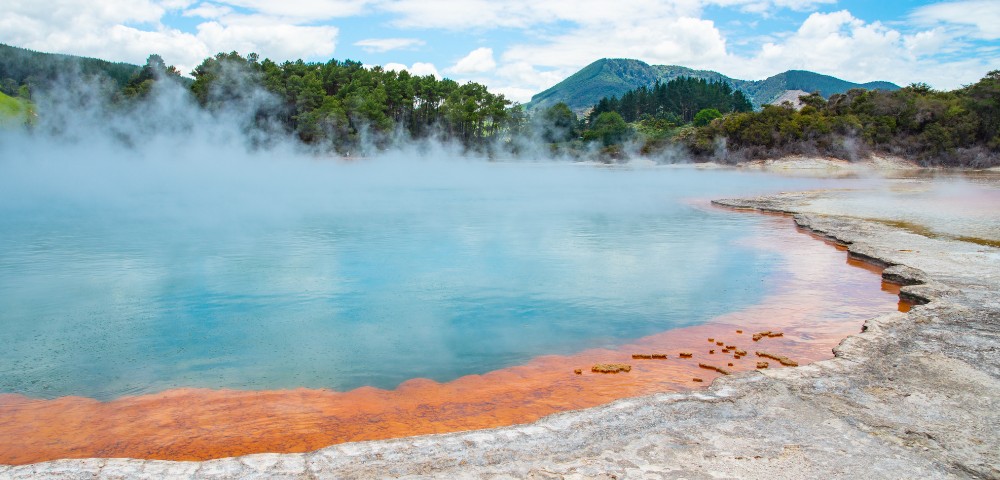 Steaming geothermal pool with vibrant orange mineral deposits at the edge, bordered by forested hills and a clear blue sky in the background.
