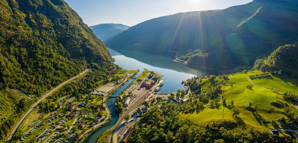 Aerial view of a scenic fjord with sunlit mountains, lush green fields, and a small river flowing through a village, conveying tranquility and beauty.