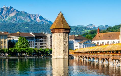Scenic view of Chapel Bridge in Lucerne with its iconic tower, flanked by historic buildings. Snow-capped mountains rise majestically in the background.