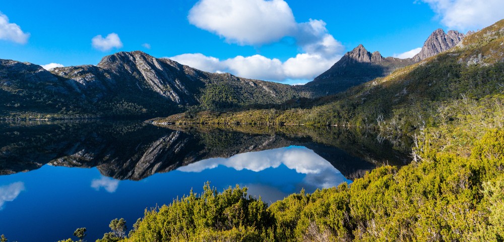 A serene mountain landscape with clear blue skies and fluffy clouds reflects perfectly on a still lake. Greenery in the foreground adds vibrant contrast.