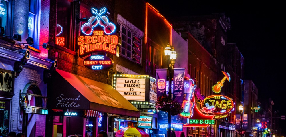 Bright neon lights illuminate a lively Nashville street at night, featuring colorful bar signs, including "The Second Fiddle" and vibrant guitar motifs.