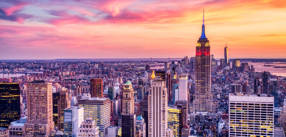 A vibrant New York City skyline at sunset, featuring towering skyscrapers with the Empire State Building prominently in the center under a colorful, glowing sky.
