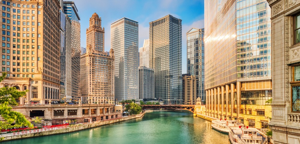 Chicago cityscape with tall, reflective skyscrapers lining a serene river, under a clear blue sky. The scene feels vibrant and bustling.