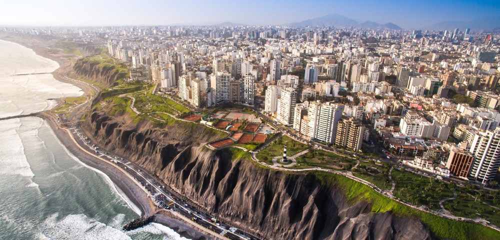 Aerial view of a coastal cityscape with tall buildings lining dramatic cliffs. Waves crash against the shore, and green parks stretch along the cliffs' edge.