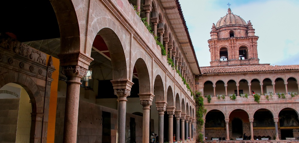 Colonial-style courtyard with arched stone colonnades leading to a large church bell tower. The atmosphere is historic and serene.