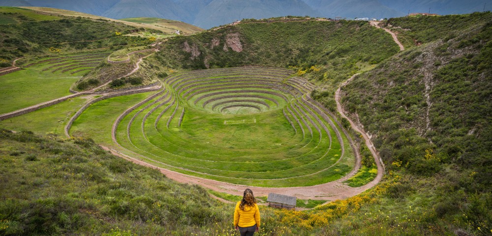 A woman in a yellow jacket stands overlooking Moray, Peru's circular terraced ruins surrounded by lush greenery and distant mountains, conveying wonder and awe.