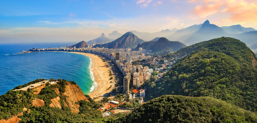 Aerial view of a vibrant coastal cityscape featuring a sprawling beach lined by urban buildings, flanked by lush green mountains under a colorful sky.