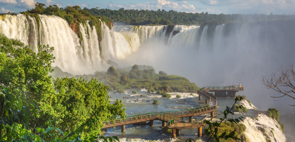 Scenic view of Iguazu Falls, with cascading waterfalls surrounded by lush green foliage. A winding wooden walkway extends over the misty river below.