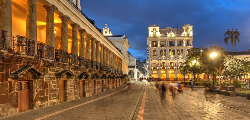 Historic plaza at dusk with illuminated colonial architecture, stone walkway, and blurred figures conveying motion. A palm tree adds to the serene ambiance.