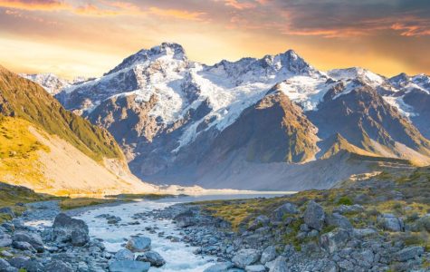 A breathtaking view of snow-capped mountains under a vibrant sunset sky, with a river flowing through a rocky valley, conveying tranquility and awe.