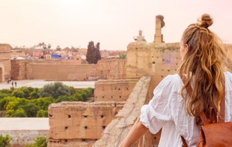 A woman with long hair and a brown backpack stands on an ancient stone wall, overlooking a historic cityscape at sunset, conveying a sense of wonder and exploration.