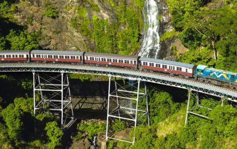 A train crosses a metal bridge over lush greenery, with a cascading waterfall in the background, conveying a sense of adventure and natural beauty.