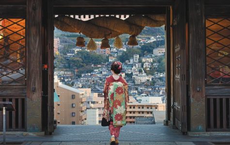 A person in a traditional Japanese kimono stands with their back to the viewer, looking out over a hillside city from within a large wooden gate adorned with a thick rope and straw ornaments.