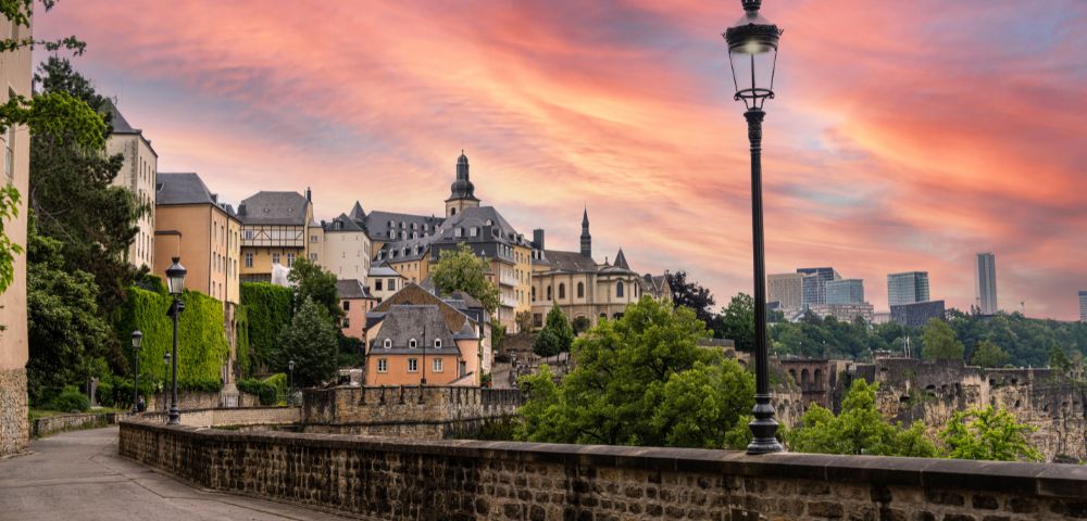 Sunset over Luxembourg city with historic buildings, lush greenery, and modern skyscrapers under a vibrant pink and orange sky.