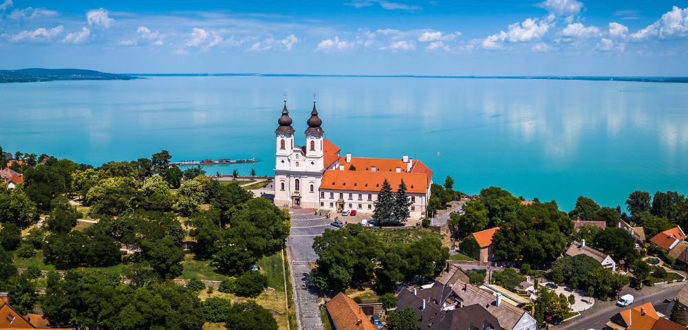 Aerial view of a picturesque lakeside village with a prominent white building featuring twin spires and a red roof, surrounded by lush greenery and calm blue water.