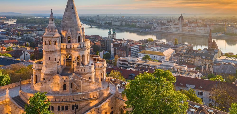 Scenic view of Fisherman's Bastion at sunset, overlooking the Danube River and Budapest city skyline. The scene is peaceful with warm golden tones.