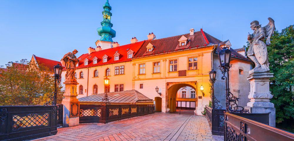 A historic European gate with a vibrant turquoise spire and red-roofed building at dusk. Statues and ornate lamps line the bridge, creating a warm, welcoming atmosphere.