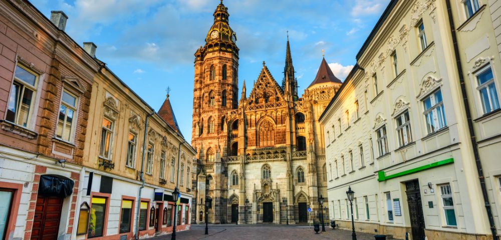 A historic Gothic cathedral towers over a cobblestone street lined with colorful 19th-century buildings, reflecting warm sunlight, evoking a serene atmosphere.