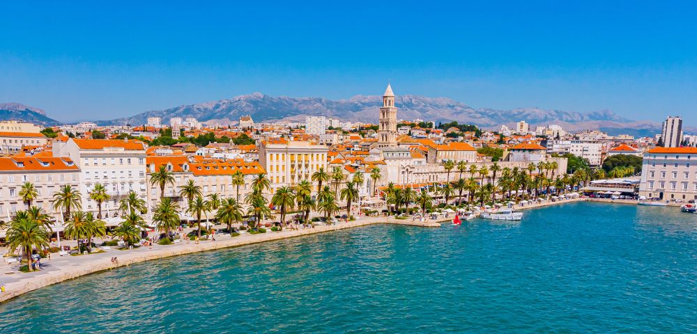 Aerial view of a coastal city with terracotta-roofed buildings, palm-lined promenade, vibrant blue sea, and distant mountains under a clear blue sky.
