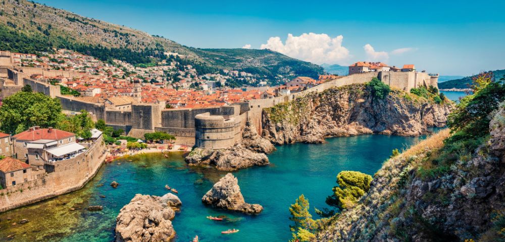 Scenic view of Dubrovnik's ancient city walls and red-roofed buildings overlooking the vibrant blue Adriatic Sea, with kayakers below and lush greenery.