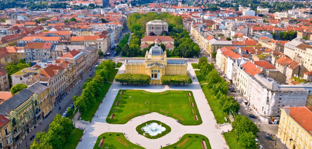 Aerial view of a historic cityscape featuring a symmetrical garden with a central fountain, bordered by tree-lined paths and surrounded by red-roofed buildings.
