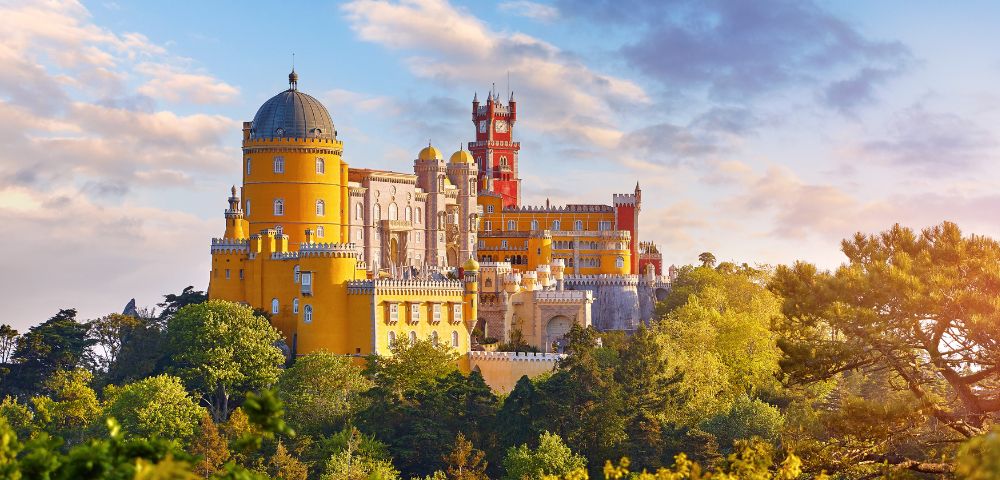 Bright, colorful hilltop castle with round towers and intricate details, set against a clear sky and surrounded by lush greenery, exuding grandeur.