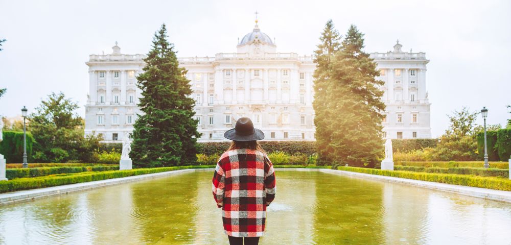 Woman in a plaid coat and hat stands by a reflective pool, facing a grand palace with soft sunlight. Tall trees flank the tranquil scene.