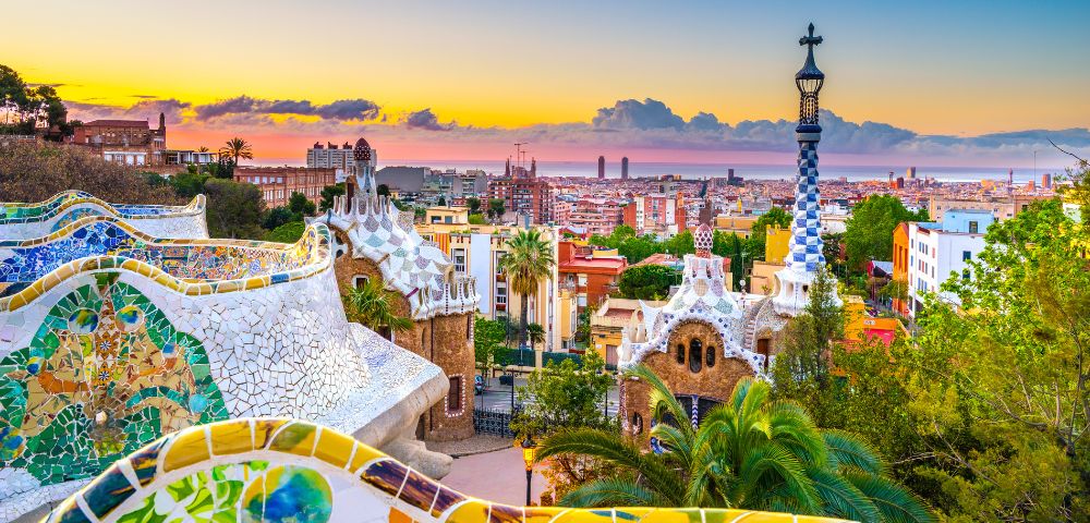 Vibrant sunset over Park Güell in Barcelona. Foreground features colorful mosaics and unique architecture, with the cityscape in the background.