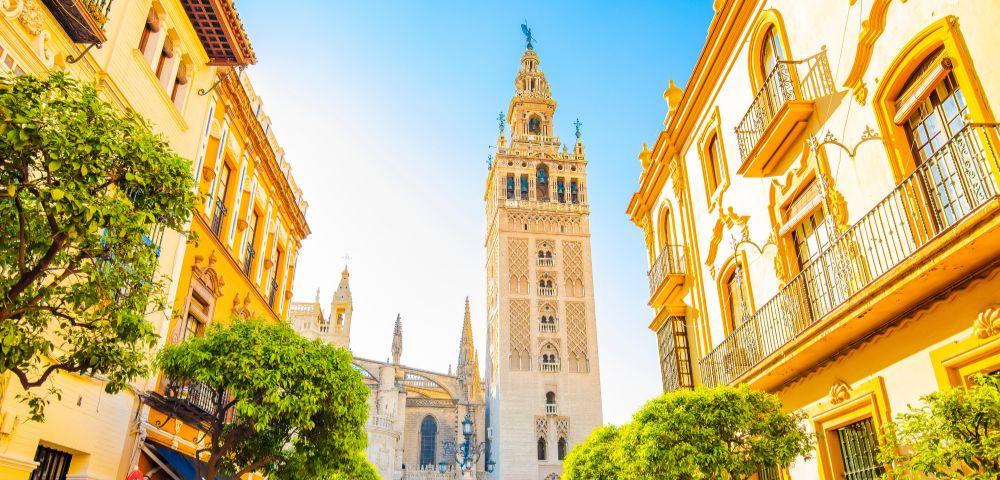 Sunlit view of La Giralda tower in Seville, Spain, framed by yellow buildings and green trees under a clear blue sky.