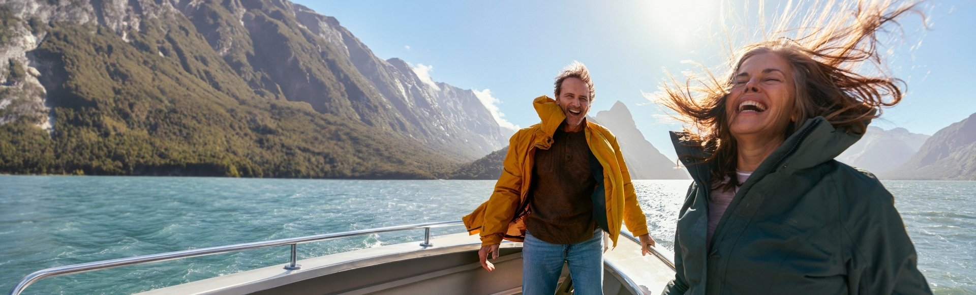 A joyful couple laughs aboard a boat on a turquoise alpine lake, with towering mountains and sunlight behind them; a carefree, adventurous moment.