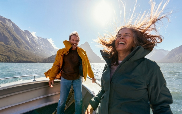 A joyful couple laughs aboard a boat on a turquoise alpine lake, with towering mountains and sunlight behind them; a carefree, adventurous moment.
