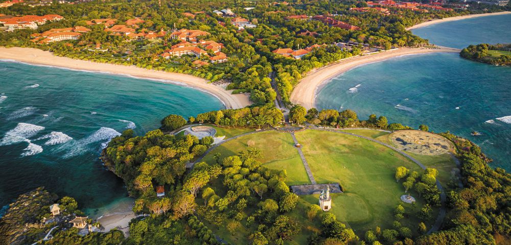 Aerial view of a lush coastal landscape, featuring two curved sandy beaches separated by a green peninsula with trees and a small building.
