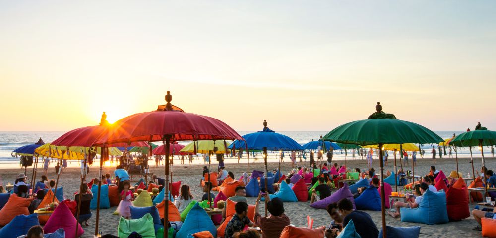 People relax on a beach at sunset, sitting on colorful bean bags under vibrant umbrellas. A relaxed, festive atmosphere with a golden sky.