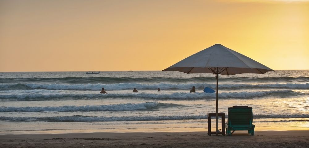 Sunset over a beach scene with gentle waves, a white umbrella, and a green lounge chair, while surfers enjoy the water nearby.