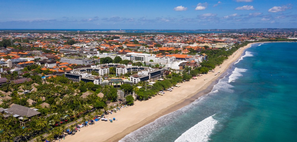 Aerial view of a sunlit beach with gentle waves and white sand. A vibrant coastal town with orange-roofed buildings lines the shore. Clear blue skies above.