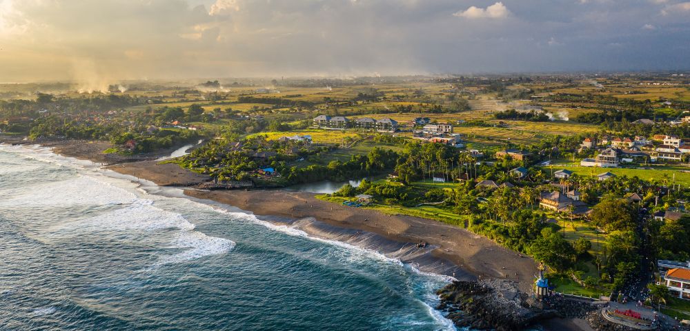 Aerial view of a coastal landscape at sunset, with waves crashing on a sandy beach. Lush greenery and scattered houses extend inland, under a cloudy sky.