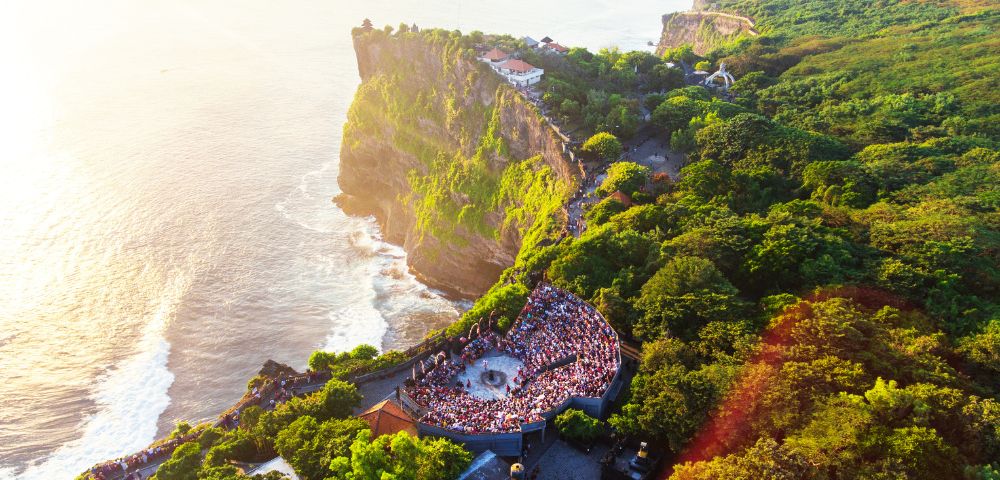 Aerial view of Uluwatu Temple, Bali, at sunset. Crowds gather in a circular area surrounded by lush greenery atop a cliff overlooking the ocean.