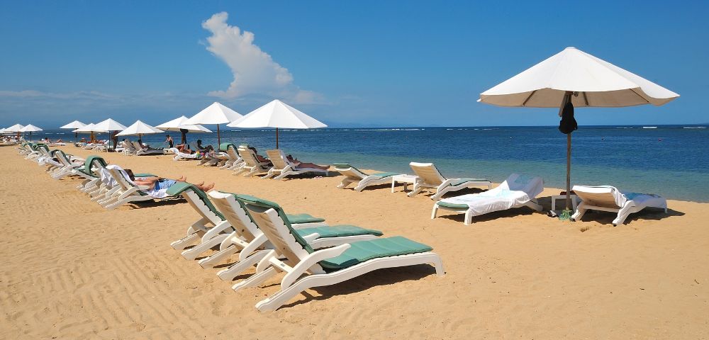 Sandy beach with rows of empty lounge chairs under white umbrellas, facing the calm blue sea. Clear sky and a peaceful, sunny atmosphere.