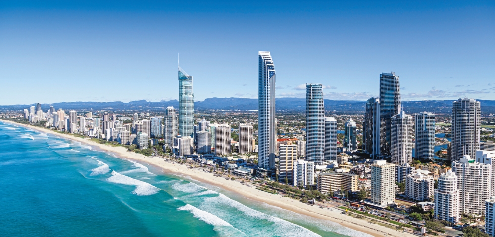 Aerial view of a coastal cityscape with modern skyscrapers lining a sunlit beach. Turquoise waves gently crash along the sandy shore under a clear blue sky.