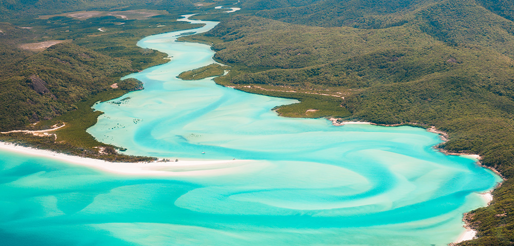 Aerial view of a winding turquoise river flowing through lush green hills, leading to a sandy beach with gentle waves.