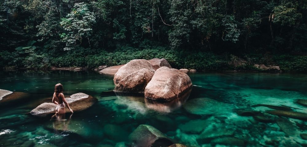 A person stands in a serene, clear turquoise river surrounded by large rocks and dense green forest. The scene conveys tranquility and natural beauty.