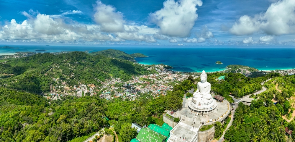 Aerial view of Phuket, Thailand, with lush green hills, a large seated Buddha statue on a hilltop, coastal town, and vivid blue ocean under partly cloudy sky.