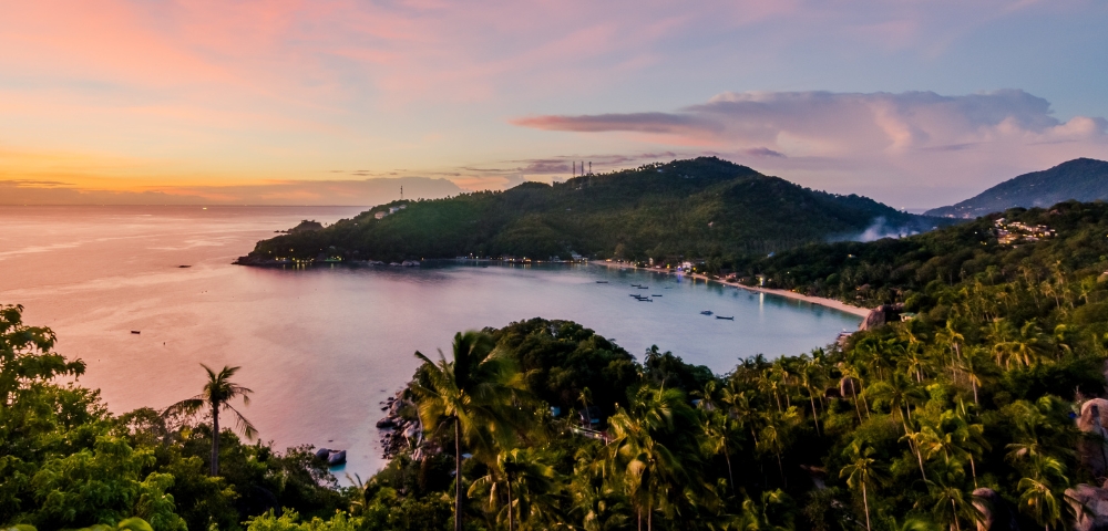 Sunset view of a tranquil bay surrounded by green hills and palm trees. The sky glows with pink and orange hues, creating a peaceful, tropical atmosphere.